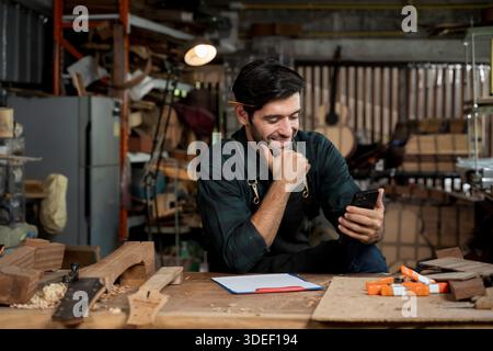 Guitar maker in apron smiles while checking a smartphone at his workbench in a woodworking shop. Artisan small business communication, customer messag Stock Photo