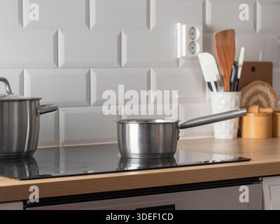 Modern kitchen counter scene with a stainless steel pot on a cooktop, cooking utensils in a ceramic holder, wooden cutting board, and white subway til Stock Photo