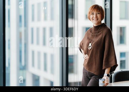 Smiling 50 year old professional woman standing near office window with city view Stock Photo