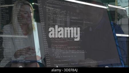 Holding notepad, colleagues in light blouse and dark blazer reviewing code on monitor in office Stock Photo