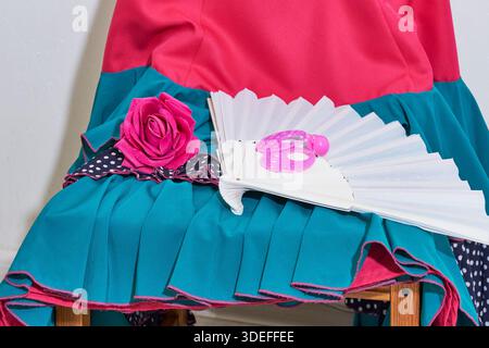Close up shot of vibrant flamenco dancer outfit in pink and teal. Folding white hand fan, bright pink hair rose, and matching earrings on chair. Stock Photo