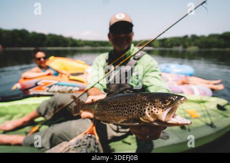 An fisherman holding a freshly caught trout while fishing from a kayak on a calm freshwater lake during summer. Stock Photo