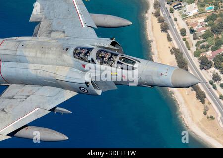 The photograph depicts a Greek Air Force F-4E Phantom II captured in flight during Athens Flying Week 2025. A close-up air-to-air angle highlights the pilot seated in the front cockpit beneath the canopy. The F-4E is a twin-seat, twin-engine fighter aircraft designed by McDonnell Douglas and powered by two General Electric J79-GE-17 turbojet engines. The type has served the Hellenic Air Force in air defense and ground attack roles and is recognizable by its swept wings, rectangular intakes, and robust airframe. Stock Photo