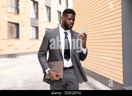 Young black CEO checking emails on cellphone, walking outdoors with laptop in hand, having break outside Stock Photo
