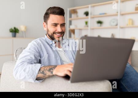 Modern job lifestyle. Happy tattooed man sitting on sofa at home and working on laptop Stock Photo
