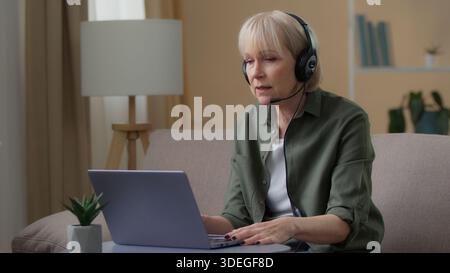 Caucasian senior woman in headset talking video call with laptop at home old female remote communicate with computer retired lady granny serious Stock Photo