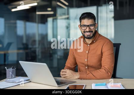 Young indian businessman with eyeglasses smiling at camera, sitting at desk with laptop in modern call center or company office, representing success and confidence in a business setting Stock Photo