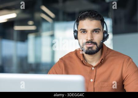 Indian man wearing a headset sitting at a desk, looking at the camera, working remotely or in a modern call center office providing client assistance and online technical support Stock Photo