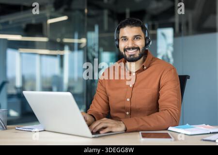 Smiling young indian customer service representative in a headset sitting at a desk, typing on a laptop and looking at the camera, providing online support Stock Photo
