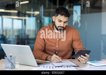 Young financial professional sitting at a desk, counting money using a calculator, reviewing documents, analyzing charts and graphs, and taking notes in a modern office environment Stock Photo