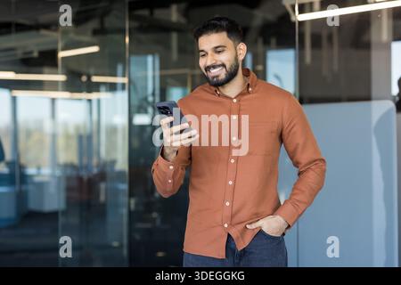 Indian man in a button-up shirt smiling and looking at his smartphone while standing in a modern office. Representing communication. Technology. And a connected lifestyle in a professional environment Stock Photo