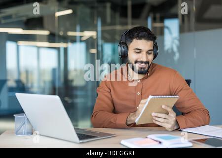 Young man wearing headphones and orange shirt actively working with laptop and notepad while smiling in a bright contemporary office environment, focusing on productivity and learning Stock Photo