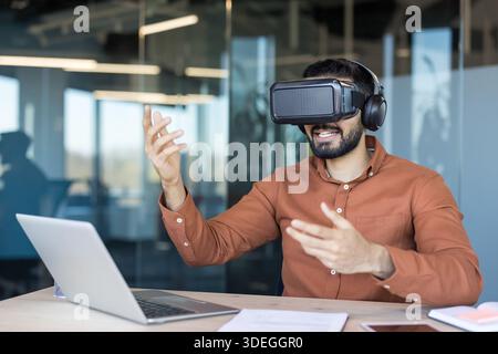 Young man wearing vr headset and headphones, interacting with virtual content while sitting at a desk with a laptop, embracing metaverse technology in a corporate environment Stock Photo