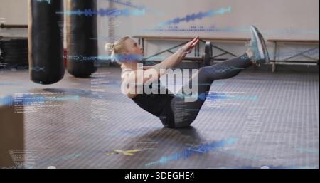 Balancing woman holding V-sit on rubber gym floor, in black workout gear, heavy bags, blue overlays Stock Photo