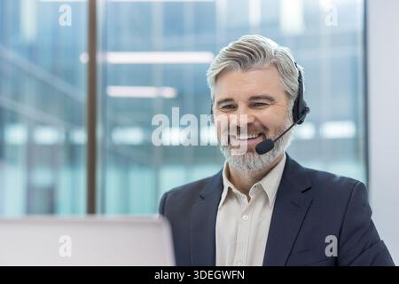 Joyful businessman close up using headset phone for video call. Mature man in business suit smiling, working inside office. Stock Photo