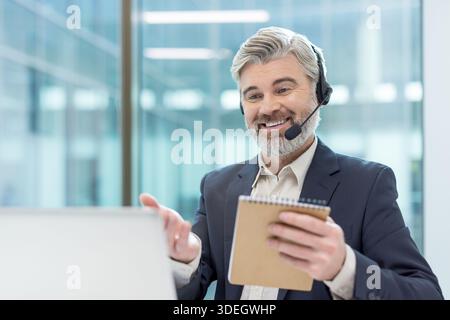 Smiling mature man wearing a headset and suit engaging in an online consultation or customer service call while holding a notebook and gesturing at a laptop screen in an office setting Stock Photo