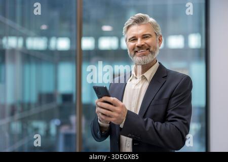 Confident mature businessman with gray hair and beard smiling at camera while holding smartphone in a modern office, portraying leadership, success and professional warmth Stock Photo