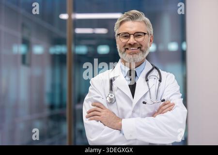 Experienced physician with gray hair and beard standing with crossed arms in a hospital corridor, representing medical care, professionalism, and trust in healthcare Stock Photo