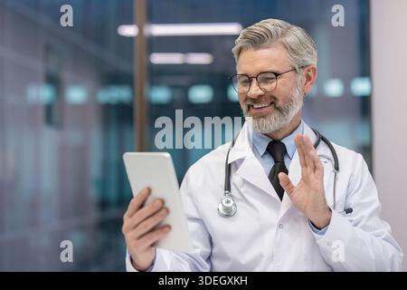 Mature doctor in white scrub uniform and stethoscope smiling, waving hand while having an online video call on a digital tablet, offering remote consultation and virtual care Stock Photo