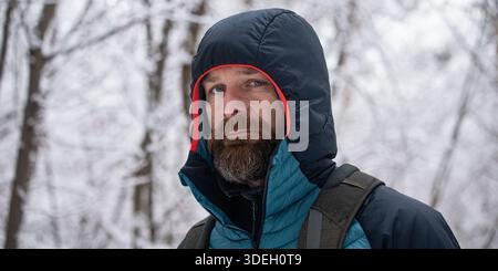 Portrait of a bearded man wearing a hooded winter jacket while hiking in a snowy forest. Outdoor adventure, cold weather, winter travel and active lif Stock Photo