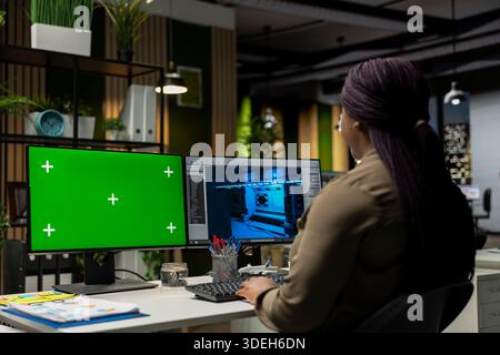 African american woman works on game developing with green screen, using dual monitors at the office. Female engineer creating a futuristic spaceship station on modern software. Stock Photo
