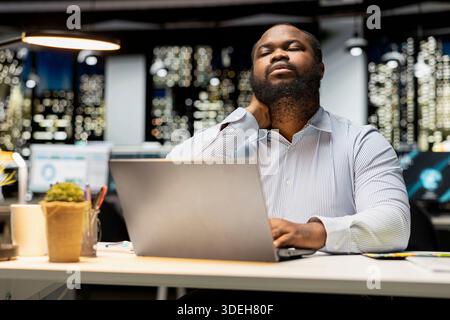 Black businessman does self massage to relieve neck stiffness and discomfort, struggling with tension due to incorrect office posture at night. Stretching his upper back muscles. Stock Photo