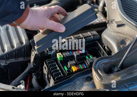 Male hand lifting plastic cover of automotive fuse box revealing colorful blade fuses and relays in car engine compartment. Concept of electrical syst Stock Photo