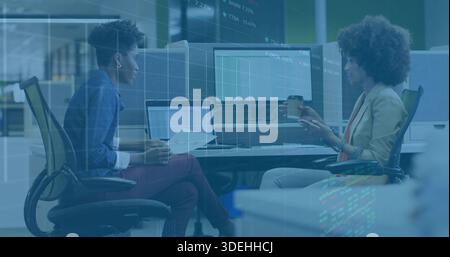Discussing printed docs, two women in denim, blazer sitting at desk with laptop, monitors and cup Stock Photo
