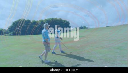 Walking senior golfers wearing blue polos and hats, carrying clubs with tee ball visible on fairway Stock Photo