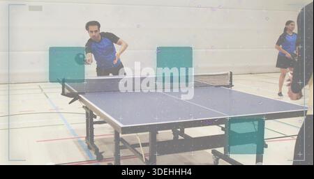 Playing left-side man wearing blue-black kit, hitting ball with paddle at table tennis table in gym Stock Photo