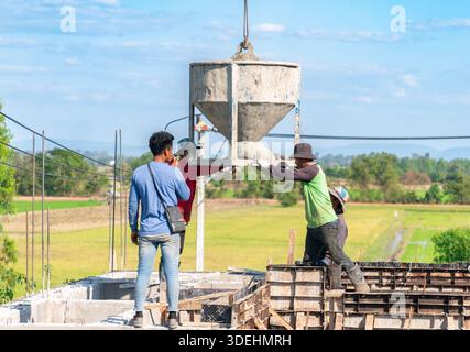 Construction workers manually guiding a large crane-lifted concrete bucket to pour wet cement into foundation formwork on a building site in a rural a Stock Photo