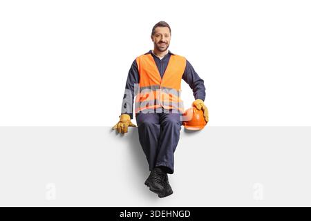 Smiling construction worker in a safety vest and hard hat sitting on a panel isolated on white background Stock Photo