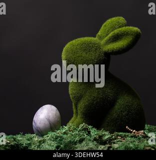 A moss-covered bunny sits next to a marbled Easter egg on a bed of moss and lichen, set against a dark background. Stock Photo