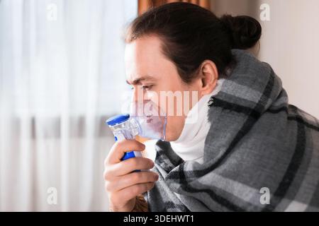 Sick man wrapped in blanket using nebulizer mask for respiratory inhalation treatment at home. High quality photo Stock Photo