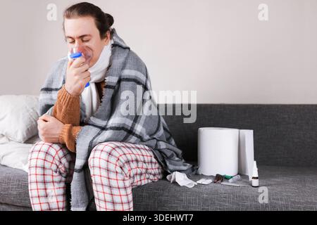 Sick man in pajamas using nebulizer mask for respiratory treatment while sitting on sofa with medicines at home. High quality photo Stock Photo