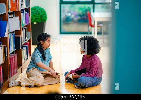 Children engaged in reading session on library floor near bookshelves. Visual conveys curiosity learning partnership diversity education development s Stock Photo