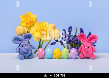 Pink and blue Easter knitted bunnies with multicolored eggs and spring muscari flowers, tulips and daffodils on the blue podium. Concept of Easter Stock Photo