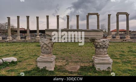 Ancient Roman columns and architectural fragments at the Agora of Smyrna, Izmir, Turkey, under a cloudy sky. Historical ruins in a grassy archaeological site. Stock Photo