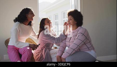 Applying center woman leaning forward, brushing makeup onto right woman at home in robe with brush Stock Photo