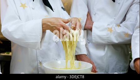 Pulling chef hands working sticky dough from white bowl in pro kitchen, white jackets black scarves Stock Photo