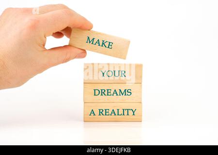 A hand carefully places a wooden block labeled MAKE atop a tower already reading YOUR DREAMS A REALITY, symbolizing building your life on solid founda Stock Photo