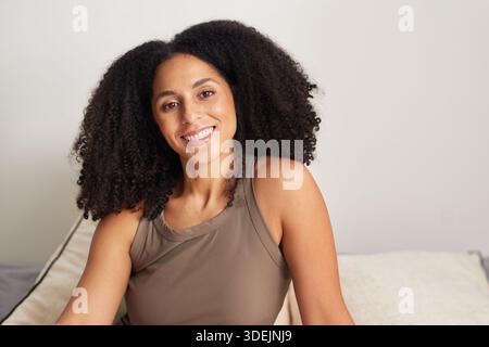 Smiling Woman With Curly Hair In Brown Tank Top Sitting On Couch At Home Stock Photo