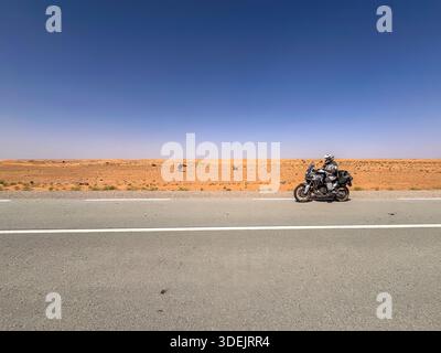Adventure motorcycle riding on a long asphalt desert road with wild camels in the background under a clear blue sky in the Sahara. Stock Photo