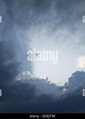 Sun rays breaking through dark storm clouds with a jet plane contrail in the sky. Stock Photo