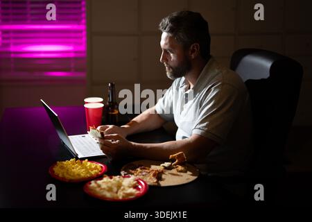 Businessman eats a late-night snack while working on laptop. A tired employee using laptop on late-night overtime. Latin Business man overworked into Stock Photo