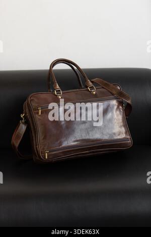 A professional brown leather briefcase with dual handles and adjustable strap sits on a sleek black leather sofa in a minimalist studio setting Stock Photo