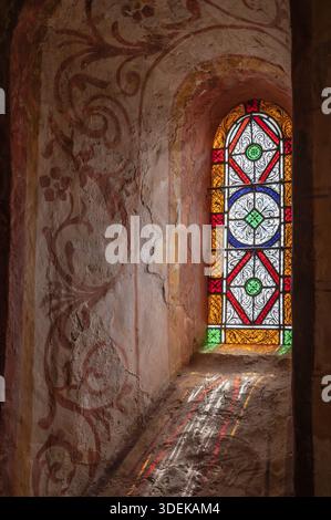 The colored lead glass of a Romanesque window opening casts colorful light reflections on the adjacent reveal with floral wall paintings in the interi Stock Photo