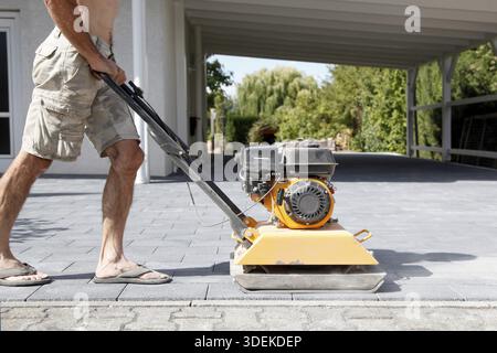 Male Worker using vibratory plate compactor to firm soil at worksite. builder. After laying of paving slabs, compacts paving stones with a gasoline vi Stock Photo
