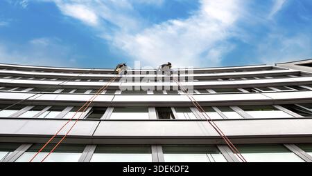 Industrial climbers wash windows of a curved building. Window cleaners cleaning glass windows on old building high in the air against blue clouds sky Stock Photo