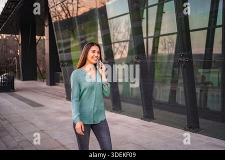 Smiling businesswoman walking near office building while talking on the phone Stock Photo